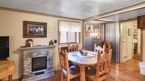 A cozy living room with a stone fireplace, wooden dining table, and chairs. A television is on the left, and there are framed pictures on the wall.