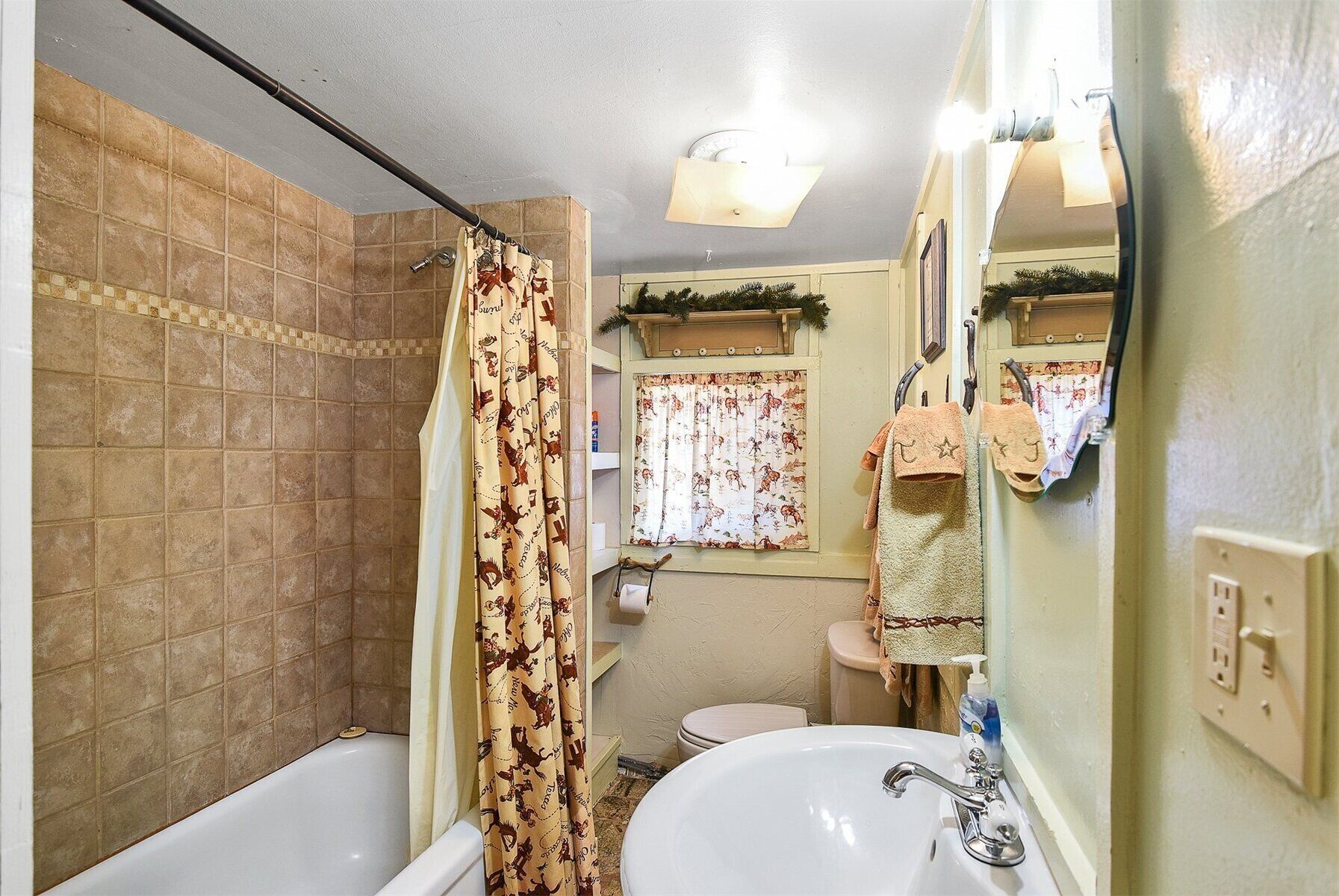 Een badkamer met een bad, wastafel en toilet. Er is een douchegordijn met een patroon, een raam met gordijnen en een spiegel.