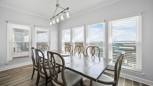 Une salle à manger avec une grande table et des chaises, de grandes fenêtres équipées de stores et un luminaire suspendu.