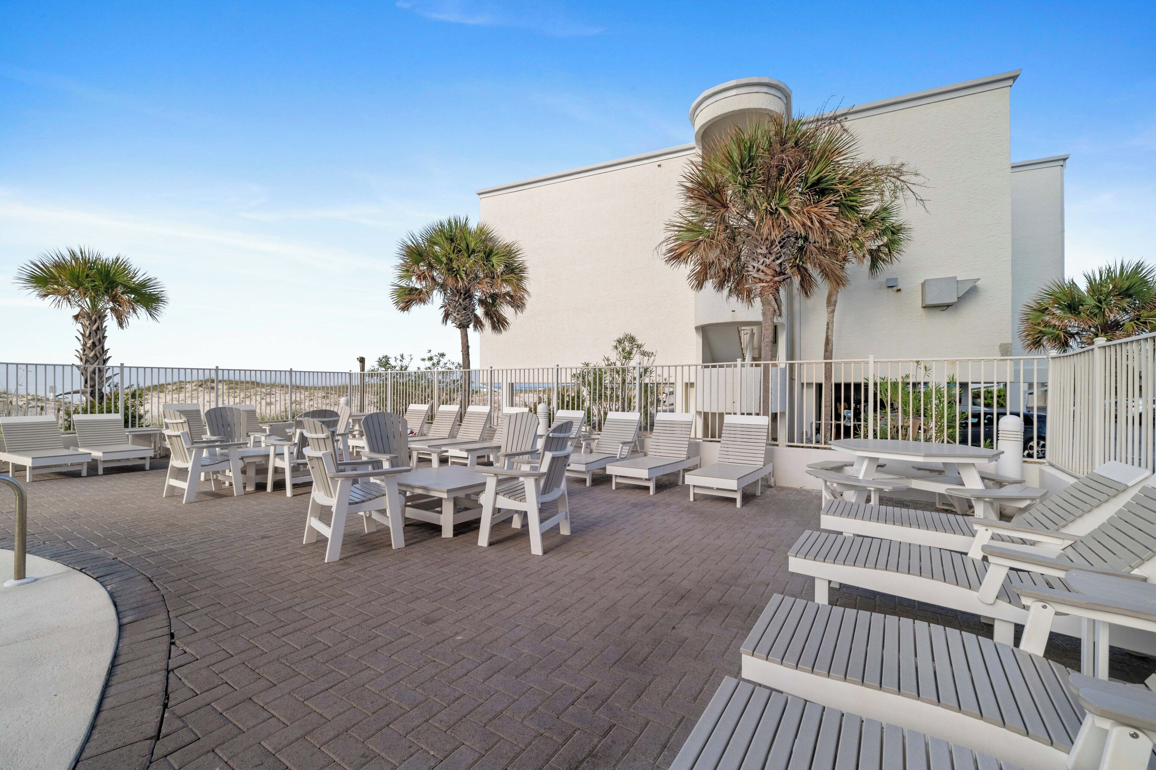 A rooftop patio with white lounge chairs and tables, palm trees, and a white building in the background.