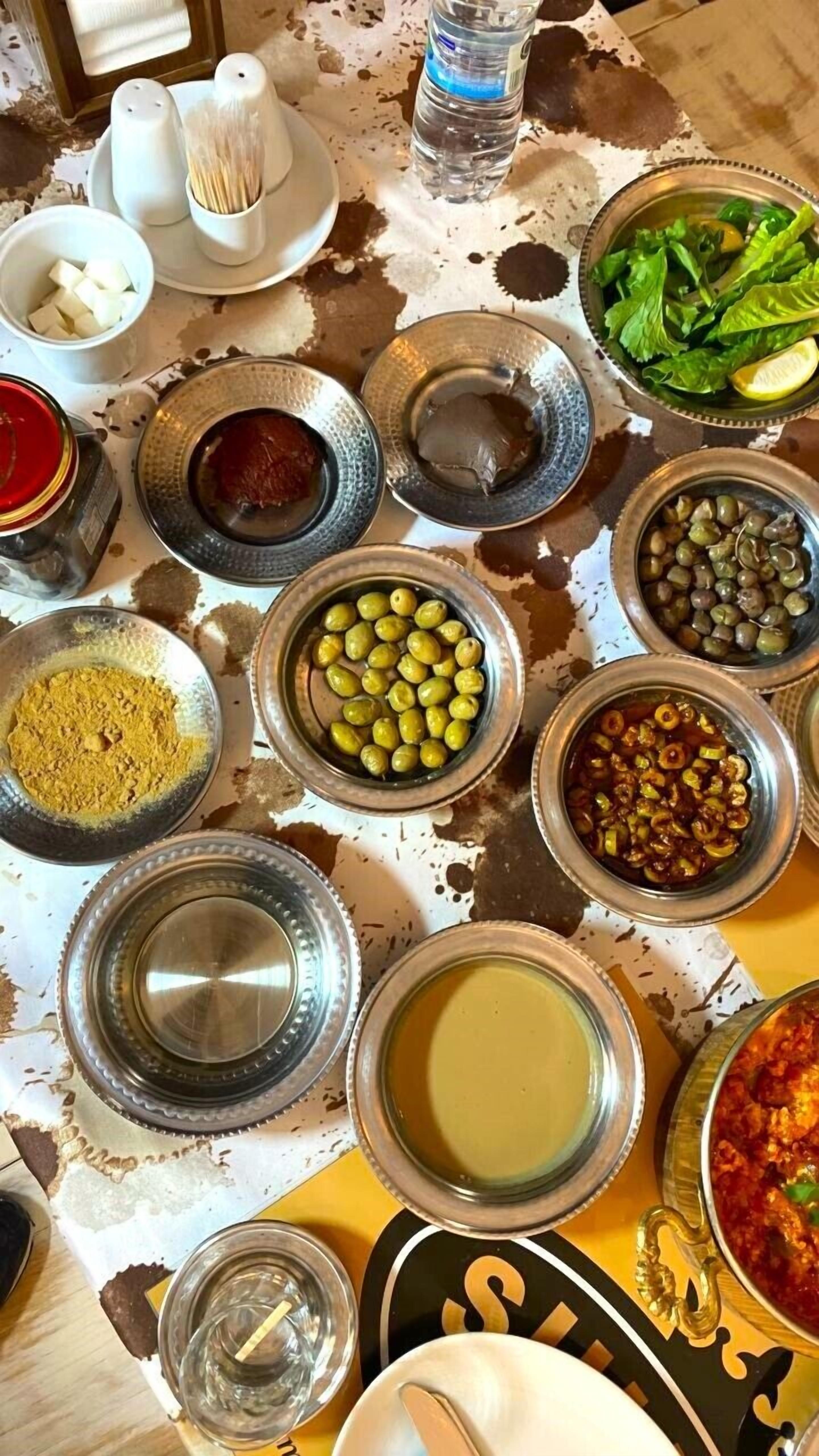 A table with various food items and condiments, including olives, pickles, and a bowl of greens.