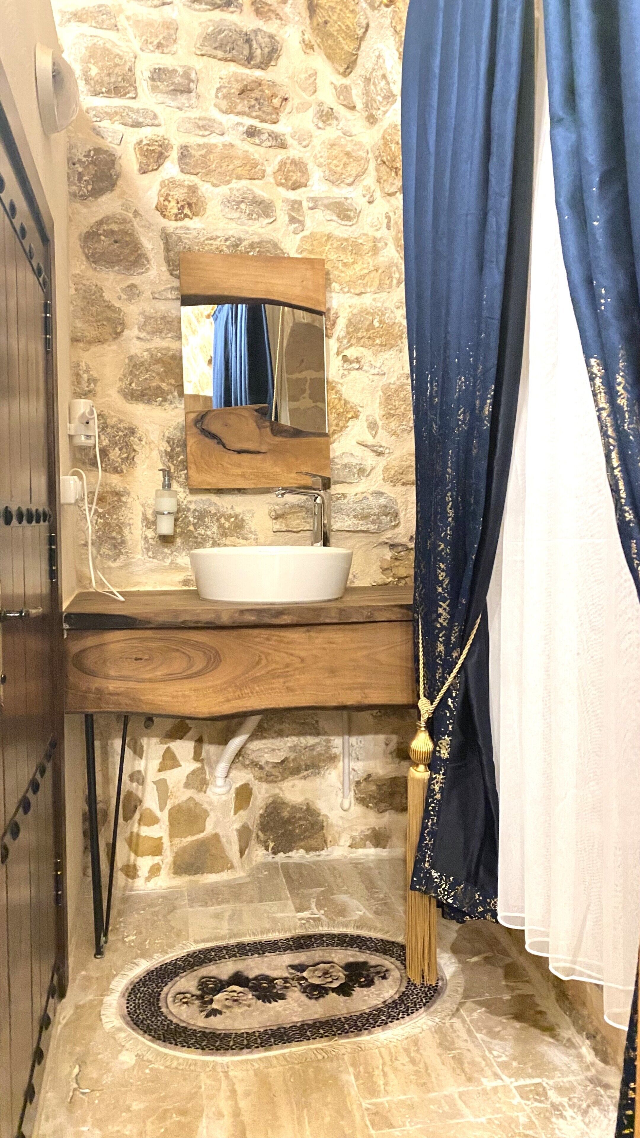 Un baño con pared de piedra, tocador de madera, lavamanos blanco, espejo, toallero y alfombra estampada.