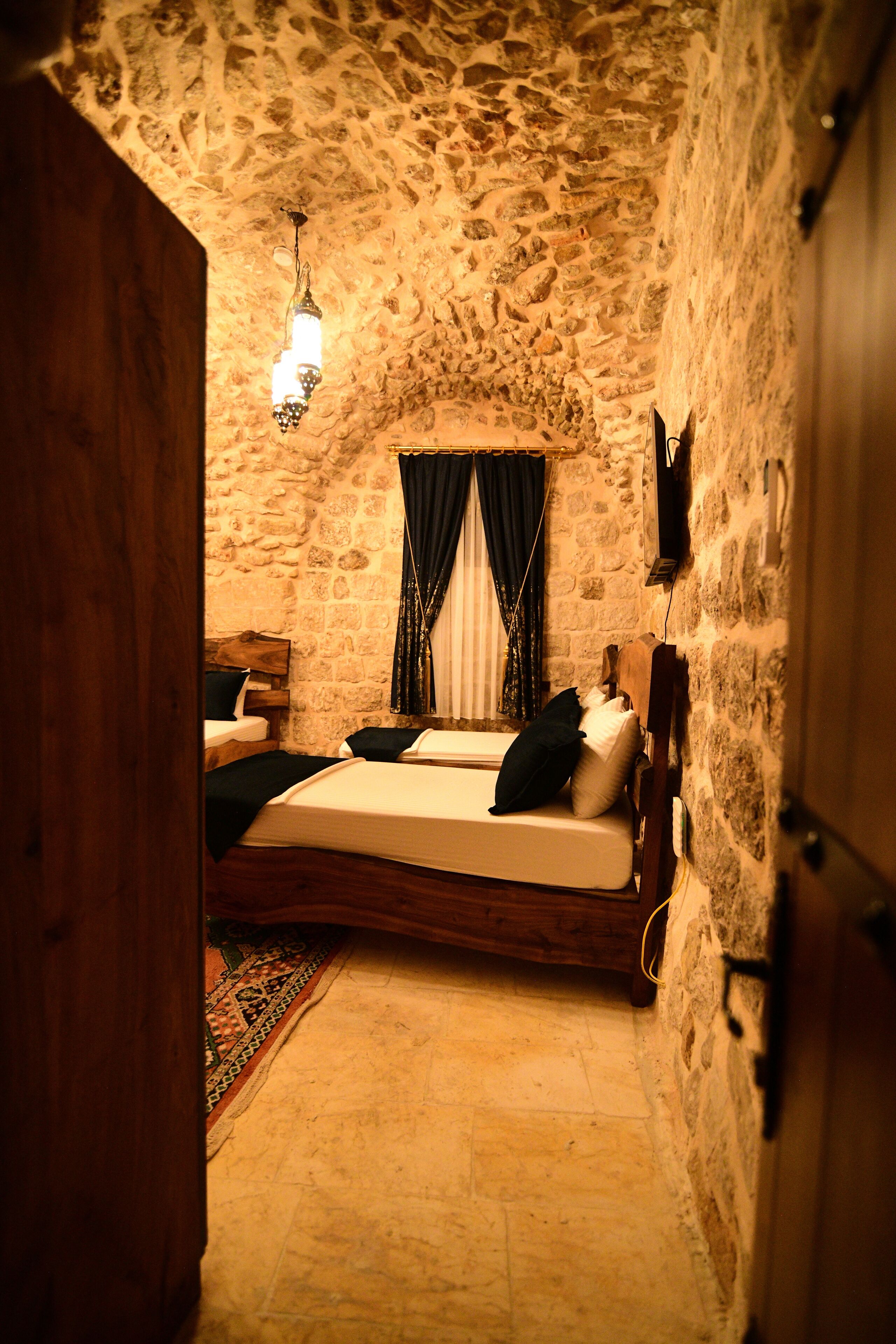 A bedroom with a stone wall, a wooden bed, a hanging light fixture, and a window with curtains.