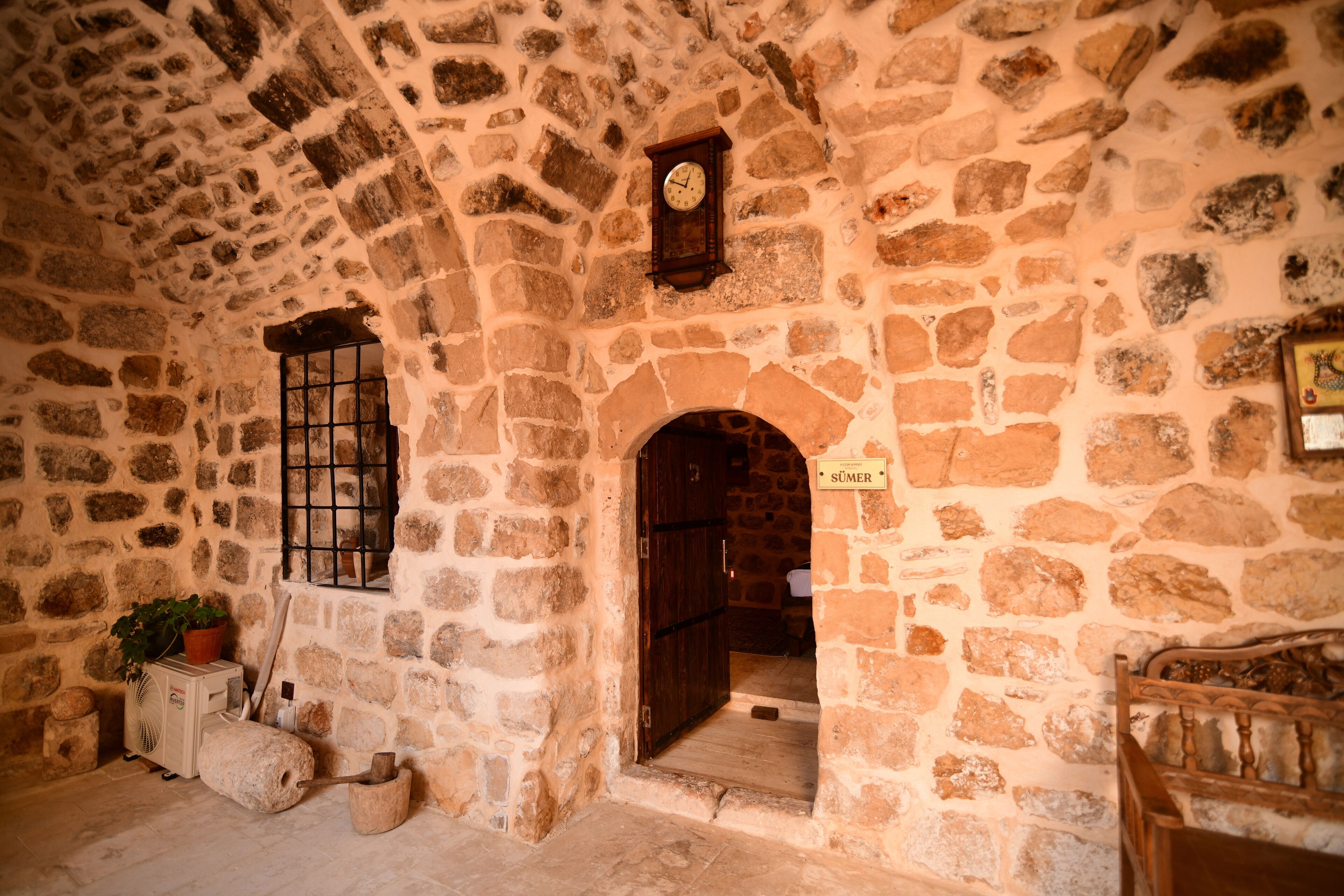 A stone-walled room with a wooden door, a window with bars, and a clock on the wall.