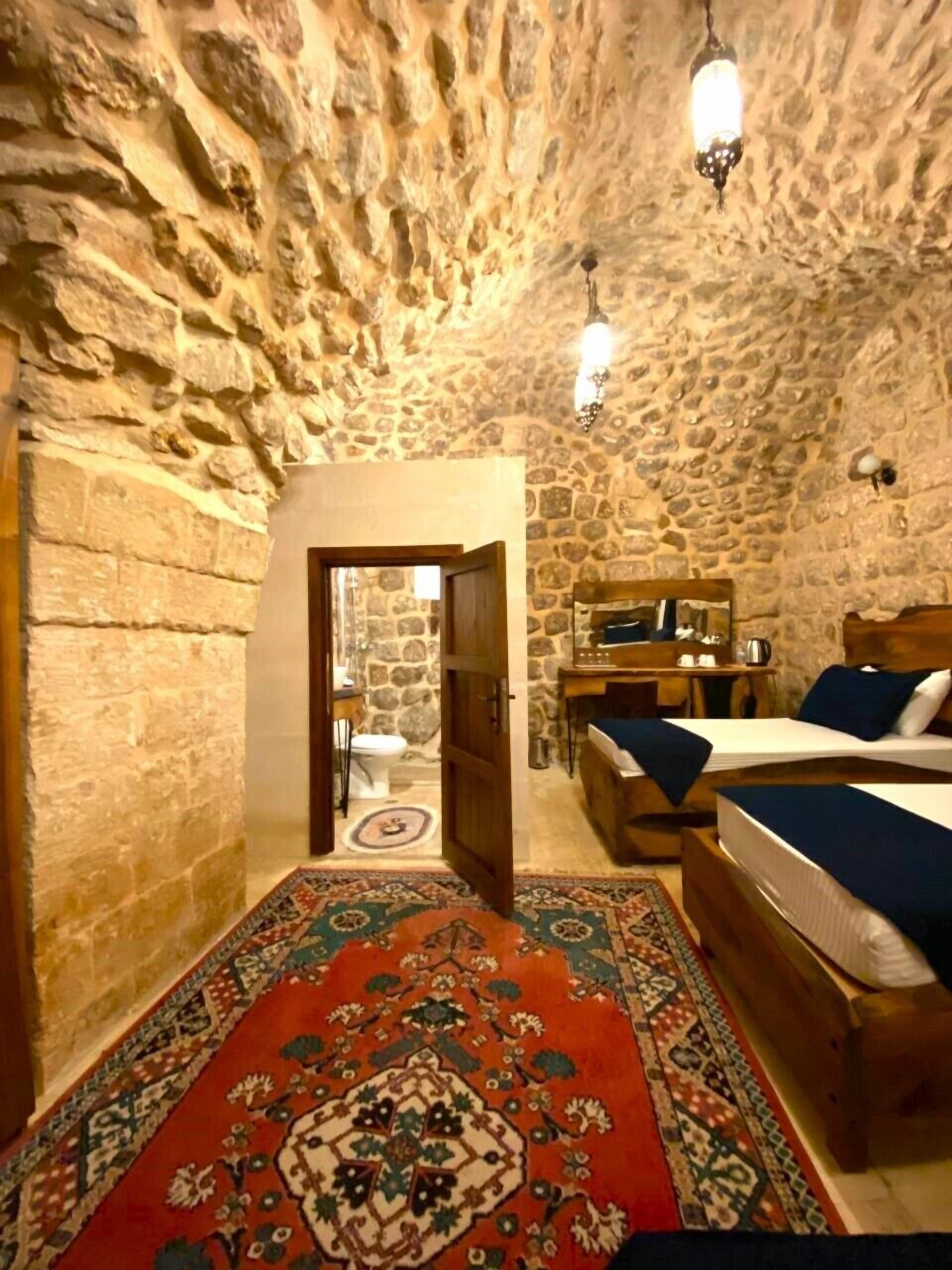 A rustic room with a stone ceiling, a wooden bed, a desk with a chair, and a patterned rug.