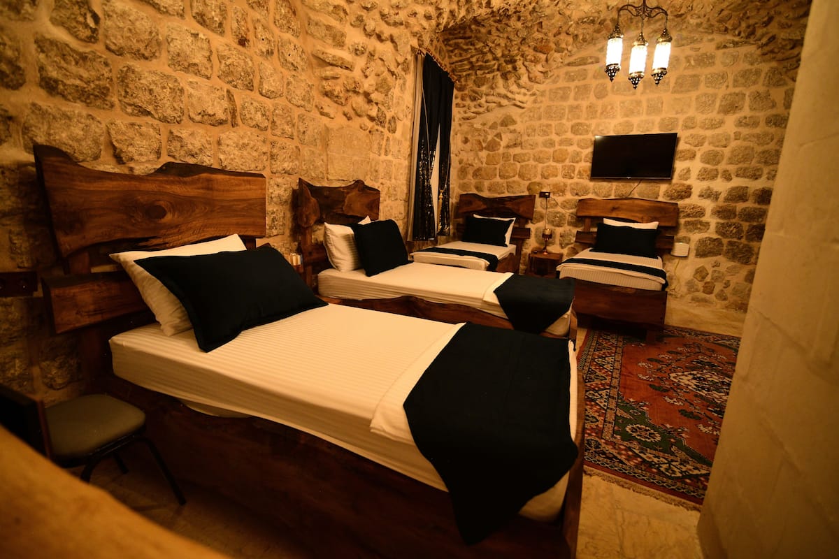 Ein Zimmer mit Steinwänden, vier Holzbetten, jeweils mit Kopfteil, einem kleinen Stuhl und einem wandmontierten Flachbildfernseher.