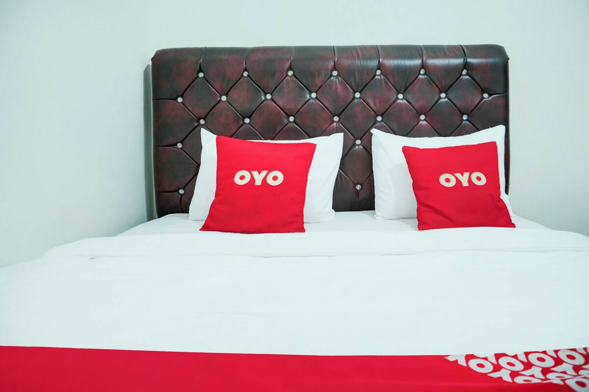 Una cama con cabecera capitonada, dos almohadas con la palabra "OYO" y una colcha blanca.