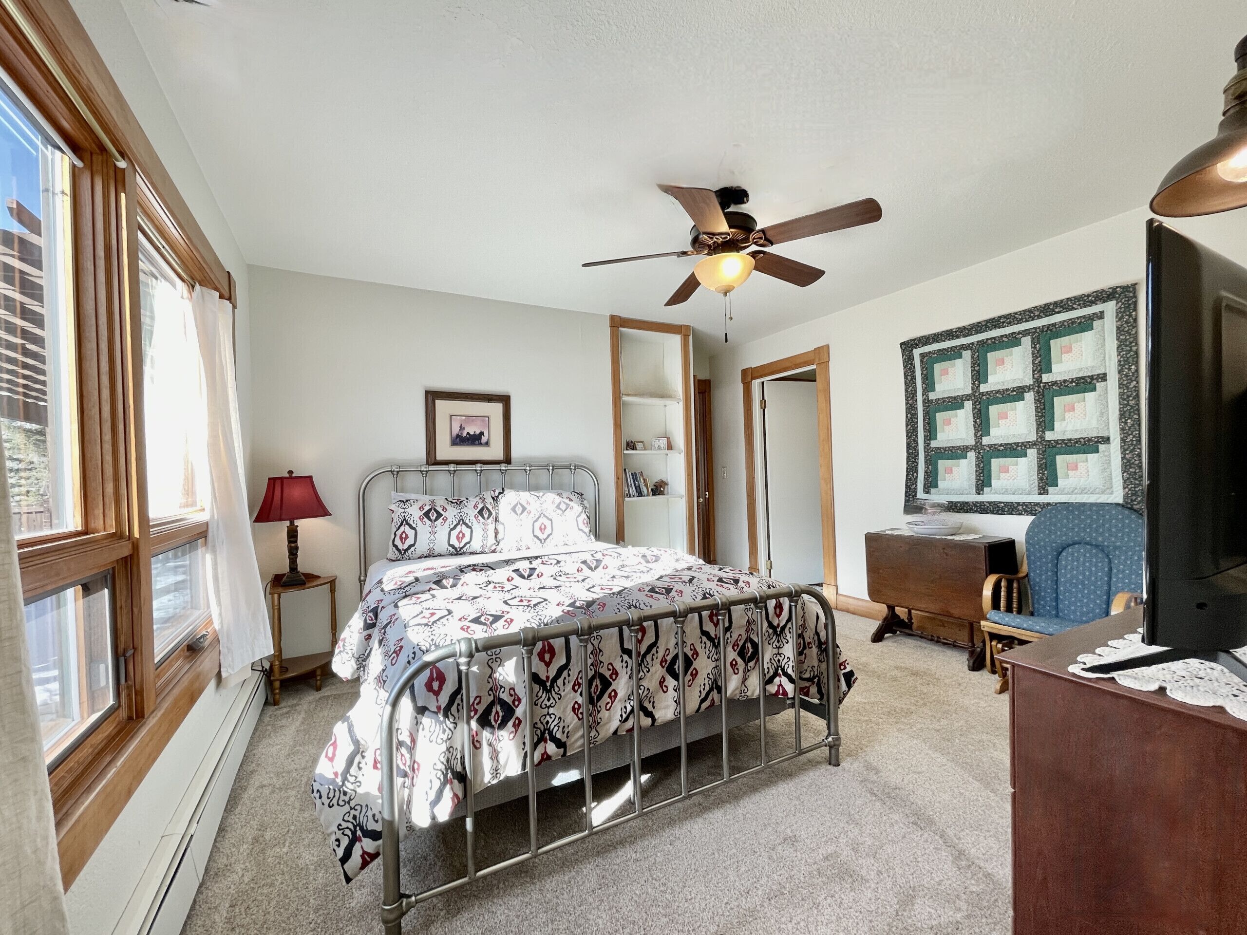 A bedroom with a bed, a desk, a chair, a ceiling fan, and a window.