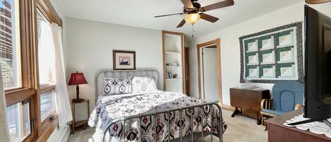 A bedroom with a bed, a desk, a chair, a ceiling fan, and a window.