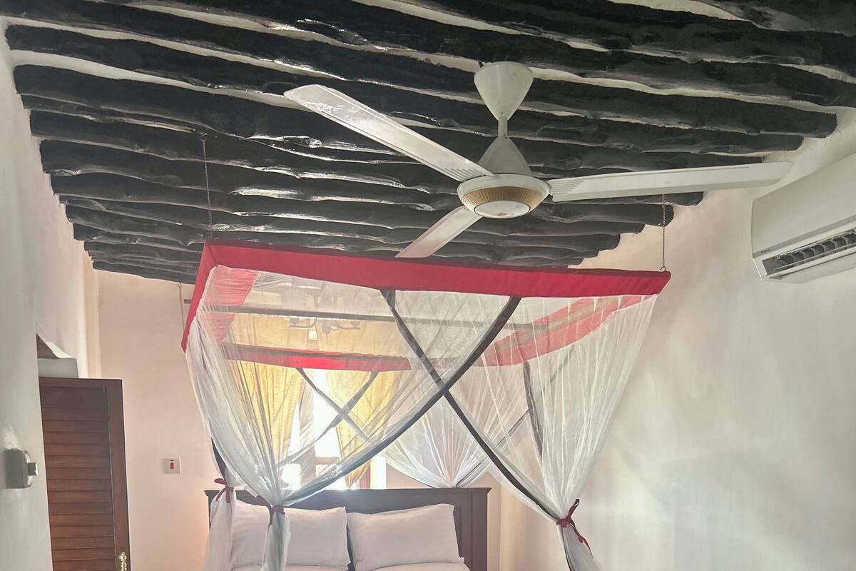 A bedroom with a bed under a mosquito net, a wooden nightstand with a kettle, cups, and a basket.