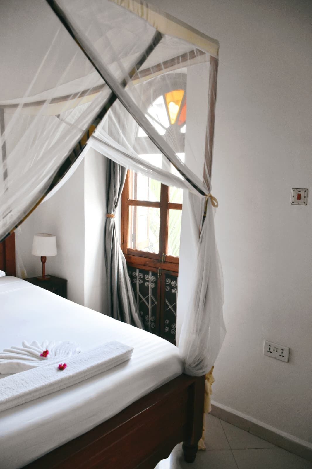 Um quarto com cama de dossel, janela com cortinas, luminária de cabeceira e uma tomada na parede.