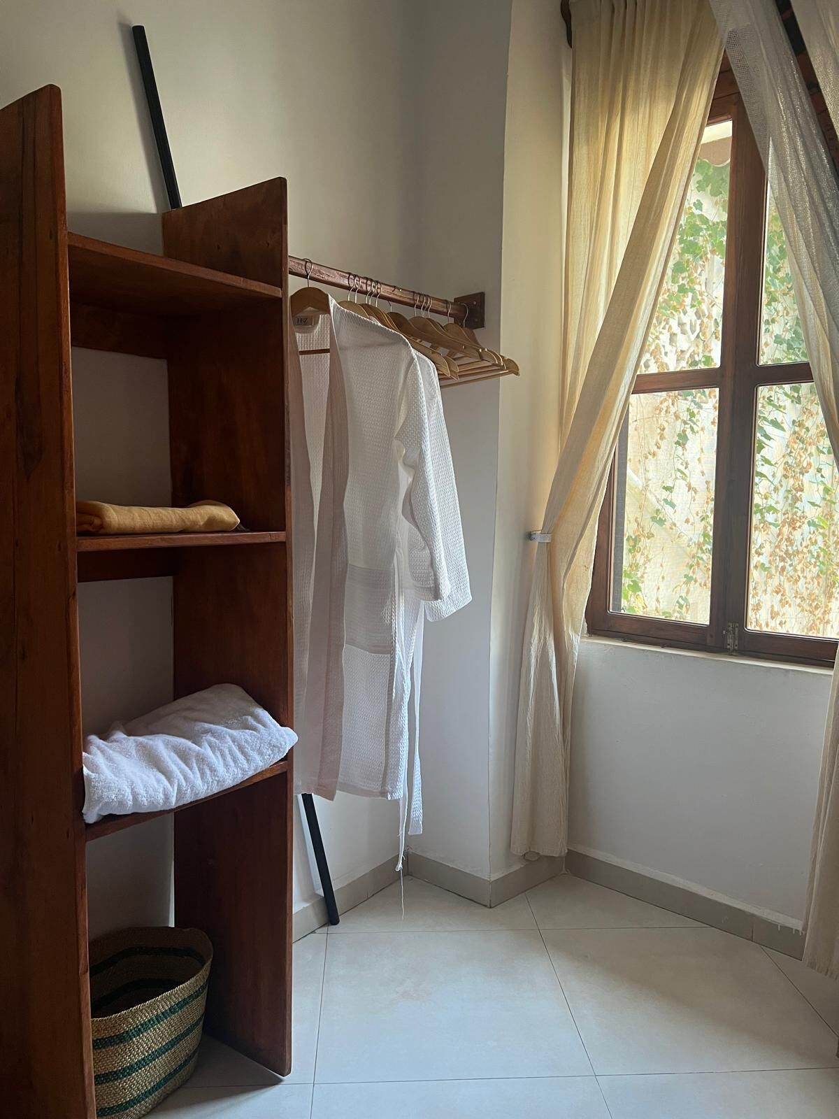 Una habitación con un armario de madera, una percha, una cesta tejida y una ventana con cortinas.