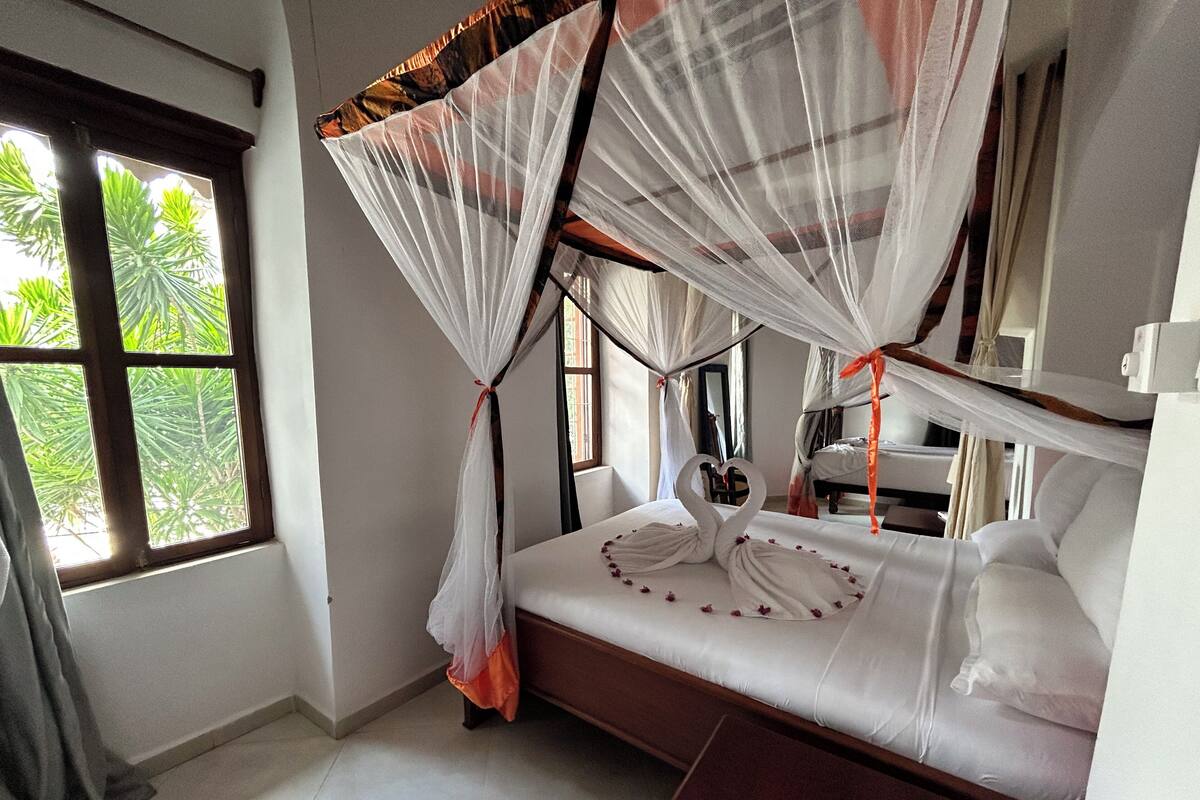 A bedroom with a canopy bed, a bedside table, and a window with a view of greenery.