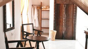 Una habitación con techo alto, una cama con cubrecama blanco, una silla de madera, una mesita pequeña y un armario con ropa colgada.