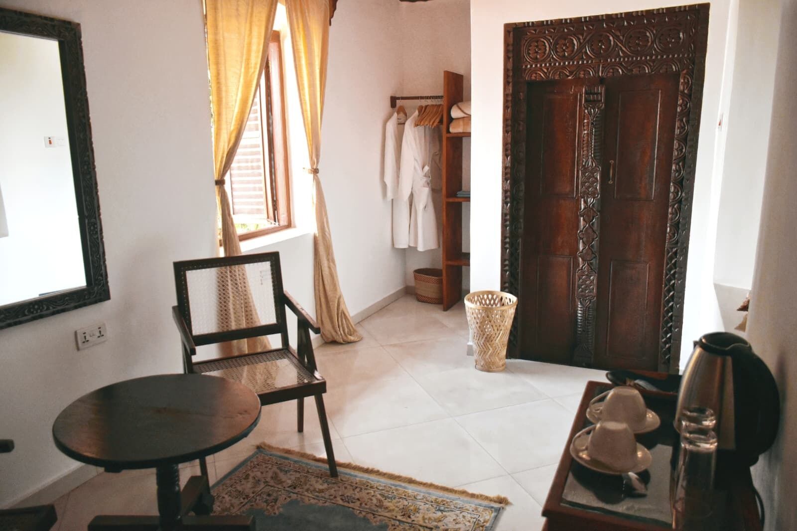 A room with a wooden door, a chair, a small table, a basket, and a window with curtains.