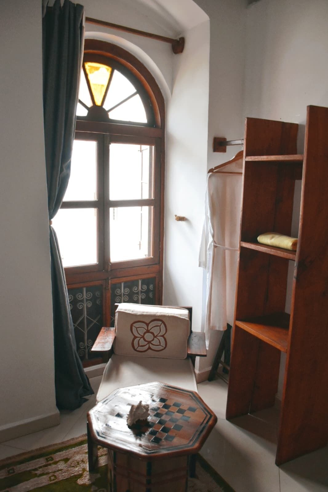 A room with a wooden bench, a small table with a chessboard, a wooden bookshelf, and a window with curtains.