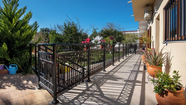 A sunlit outdoor corridor with a metal railing, potted plants, and a clear blue sky.