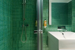 Ein Badezimmer mit grünen Fliesen, einer Dusche inklusive Duschbrause, einem weißen Waschbecken und einem Spiegel.