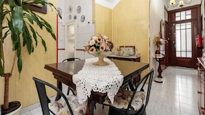 Comedor con mesa de madera, mantel de encaje blanco y un centro de mesa floral. Hay una planta en maceta a la izquierda y una lámpara de araña arriba.