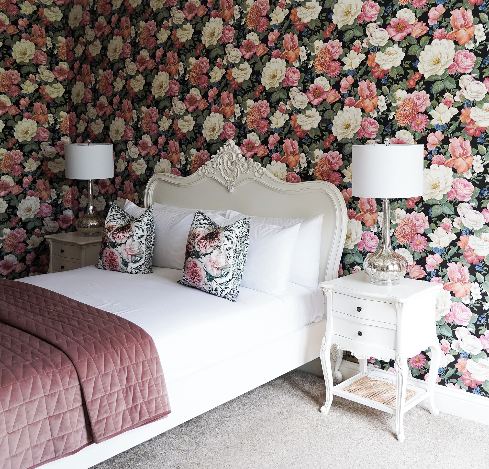 A bedroom with a floral wallpaper, a white bed with a quilted pink bedspread, two bedside tables with lamps, and two decorative pillows.