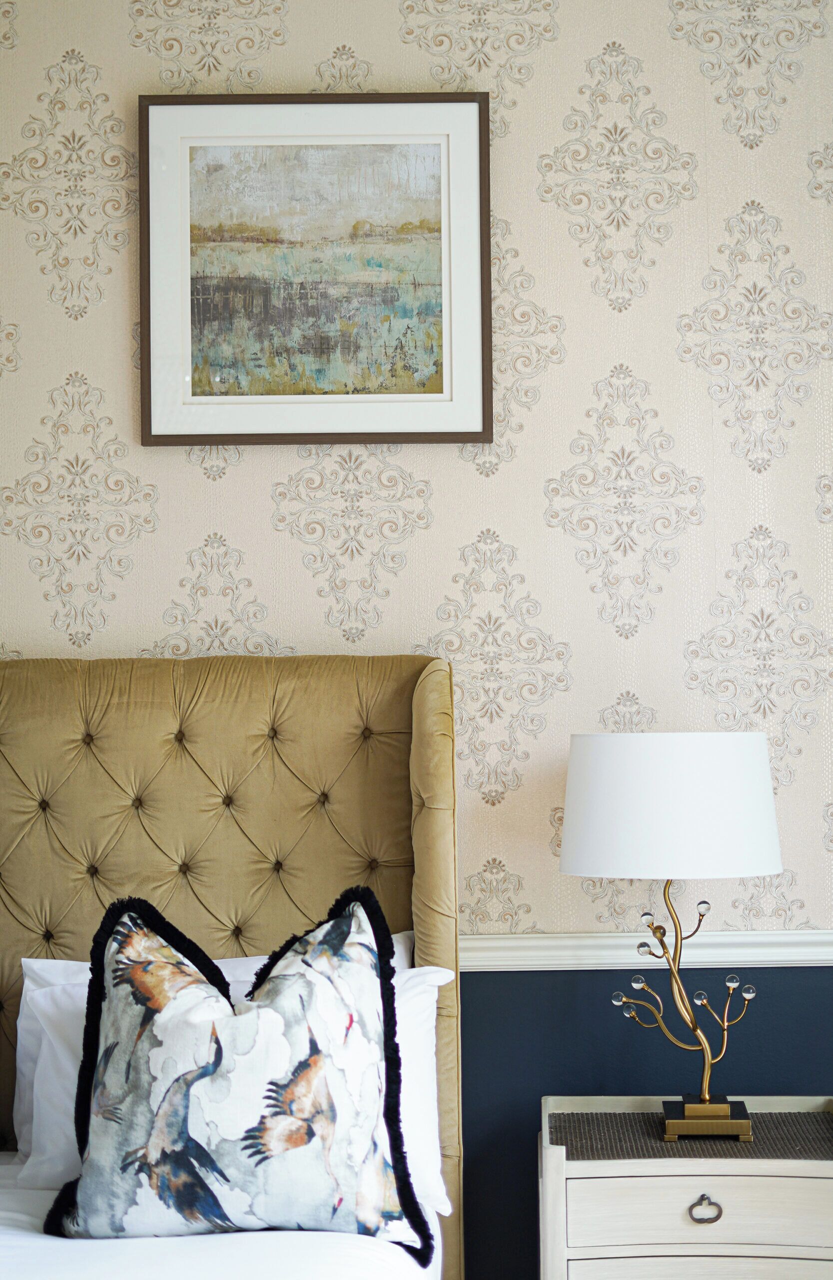 A bedroom with a tufted headboard, a framed painting, a decorative lamp, and a patterned wall.