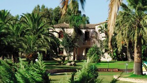 A building with a tiled roof, surrounded by lush greenery and palm trees. - Classical Residence Baia Delle Palme 2 Bedroom Apartment Sleeps 6 Persons T6 (Pula)