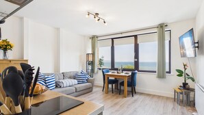 Ein modernes Wohnzimmer mit einer Couch, einem Esstisch und Stühlen, einem an der Wand befestigten Fernseher und Meerblick durch große Fenster.