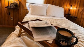 A wooden-paneled room with a bed, a bedside table with a book and a mug, a round mirror, and a window with blinds.