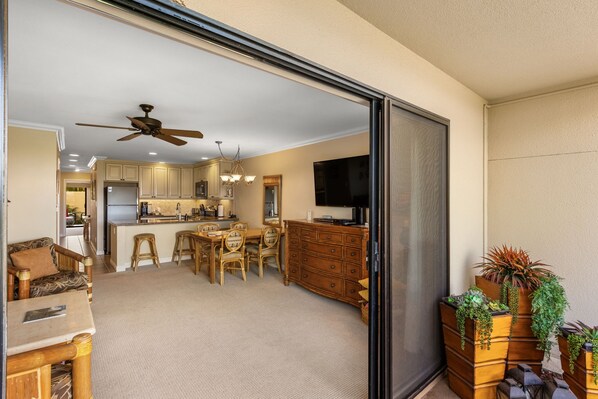 A spacious living area with a sliding glass door leading to a kitchen, a dining area with wooden furniture, and a TV mounted on the wall.