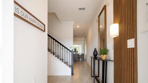 A hallway with a staircase, a sign that reads 'IS OUR HAPPY PLACE', a table with a plant, and a wall-mounted light fixture. - 2557 Awesome 4BD Townhouse Located at Storey Lake (Kissimmee)