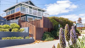 Une maison à deux étages avec un balcon, une clôture en bois et un jardin où poussent des fleurs violettes.