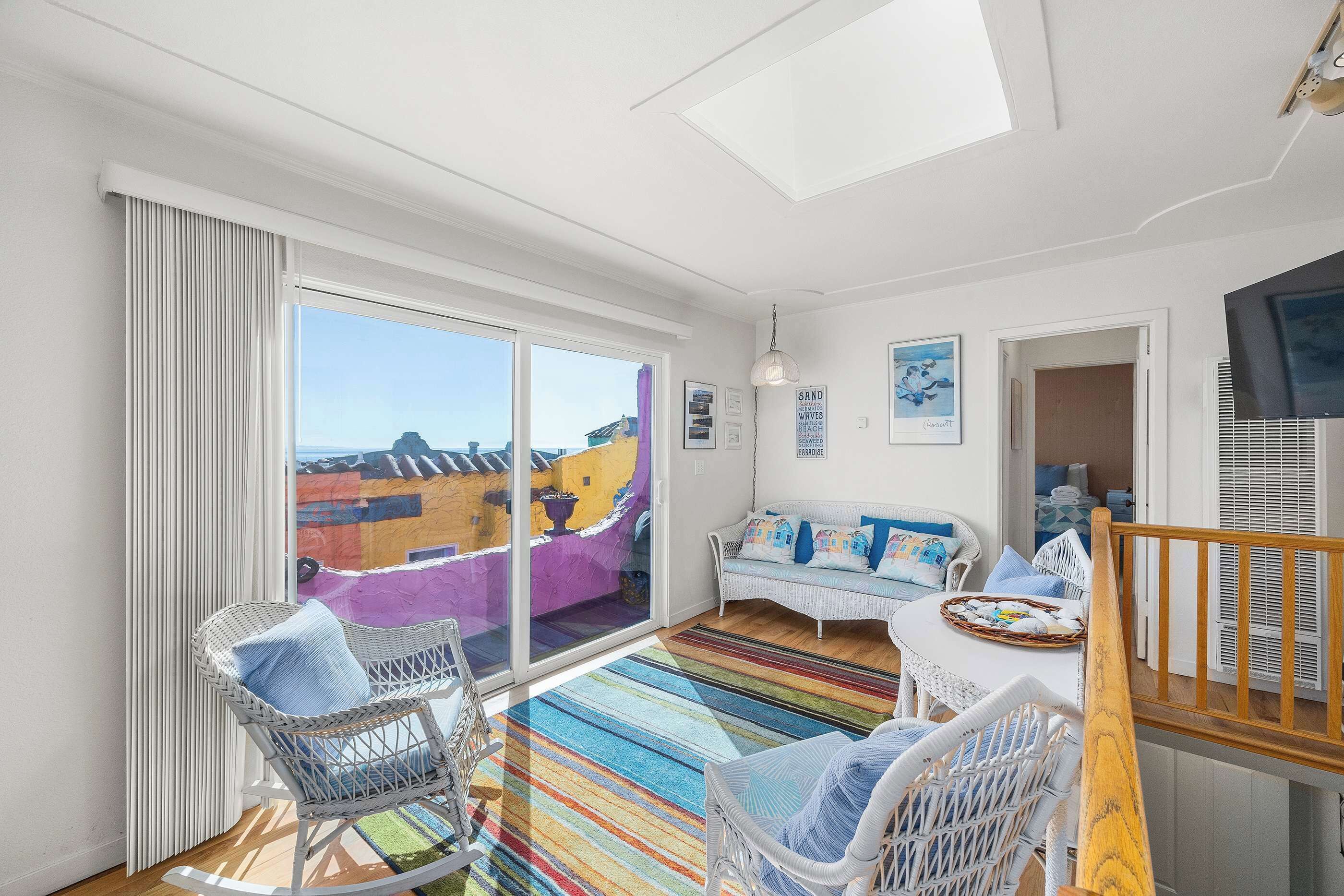 A bright room with a large window, a white sofa, wicker chairs, and a colorful striped rug.