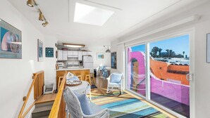 A modern kitchen with white cabinets, a dining area with wicker chairs, a colorful striped rug, and a large sliding glass door with a view of a building.
