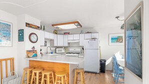 A modern kitchen with white cabinets, a central island with stools, a refrigerator, and a dining area with chairs.