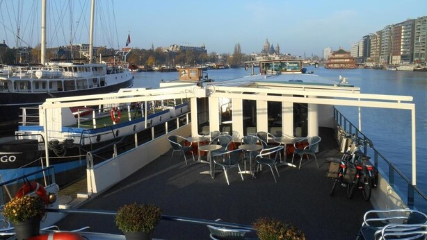 A boat deck with tables and chairs, a lifebuoy, and a view of the water and buildings in the background. - Hotelboat Iris (Amsterdam)