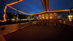 A deck with chairs and tables, overlooking a body of water with city lights in the background. - Hotelboat Iris (Amsterdam)