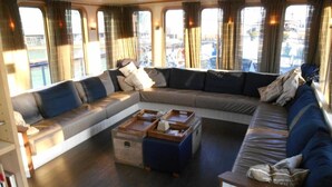 A cozy, well-lit living area with a large L-shaped sofa, a coffee table, and a view through large windows. - Hotelboat Iris (Amsterdam)