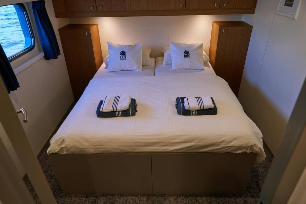 A double bed with white bedding and two pillows, a window with blue curtains, and wooden cabinets above the bed.