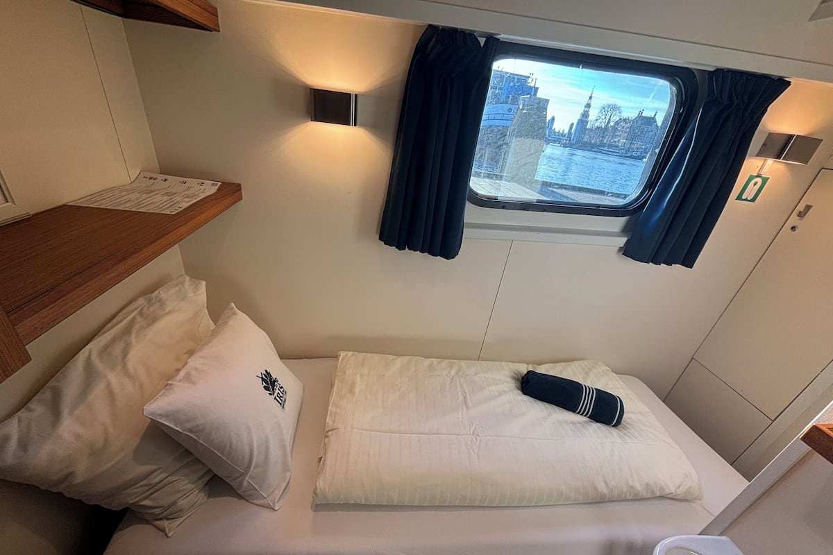 A small, cozy bed with white bedding and a single pillow, a wooden shelf with a menu, and a window with curtains.