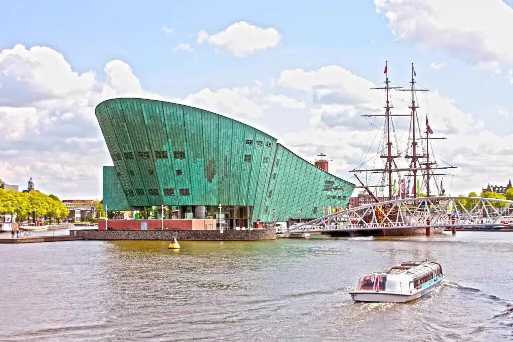 Een modern gebouw met een golvend dak, een hoge driemaster en een klein bootje op het water.