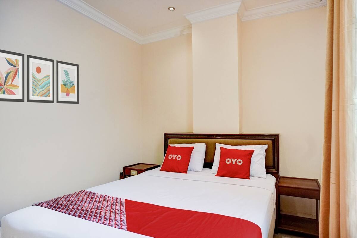 Une chambre d’hôtel avec un lit, deux oreillers portant la marque « OYO », une couvre-lit rouge et blanche, et trois œuvres d’art encadrées accrochées au mur.