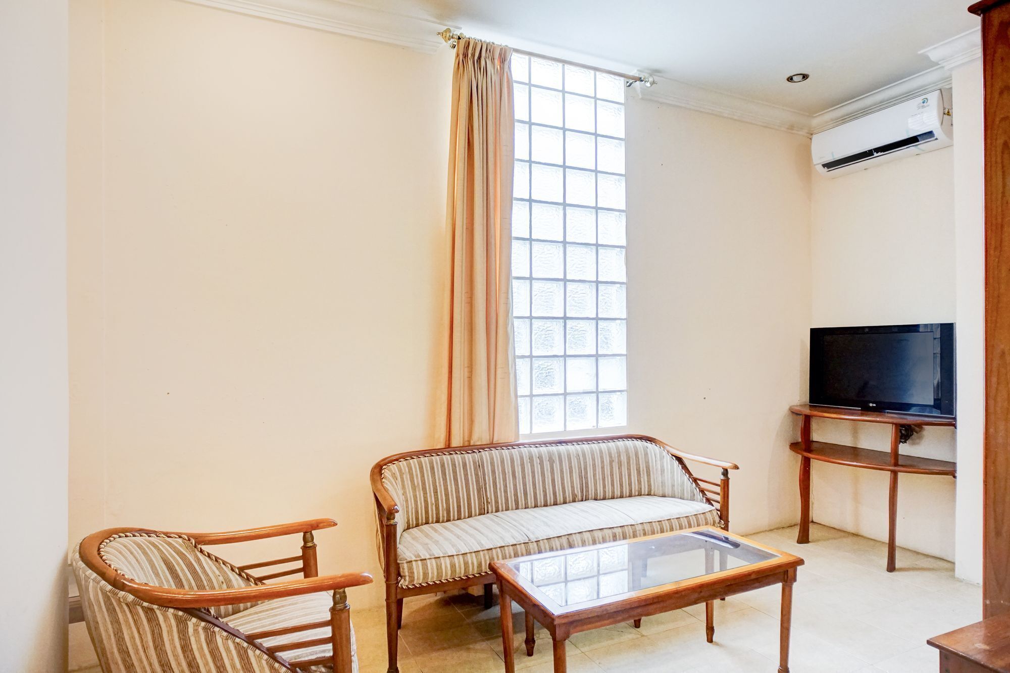 Un salon comprenant un canapé, deux fauteuils, une table basse, un téléviseur posé sur un meuble et une grande fenêtre avec des rideaux.
