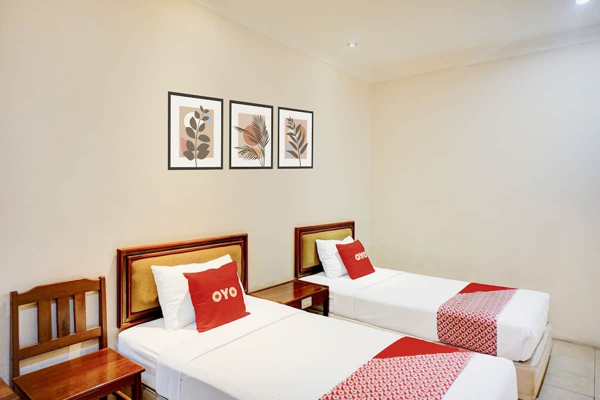 Une chambre d’hôtel avec deux lits, chacun doté d’une tête de lit en bois et d’un oreiller rouge orné du logo « OYO ». Des motifs de feuilles encadrées ornent le mur au-dessus des lits.