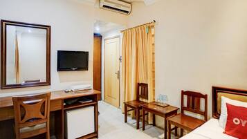 A hotel room with a desk, chair, TV, bed, and a small table with a bottle.