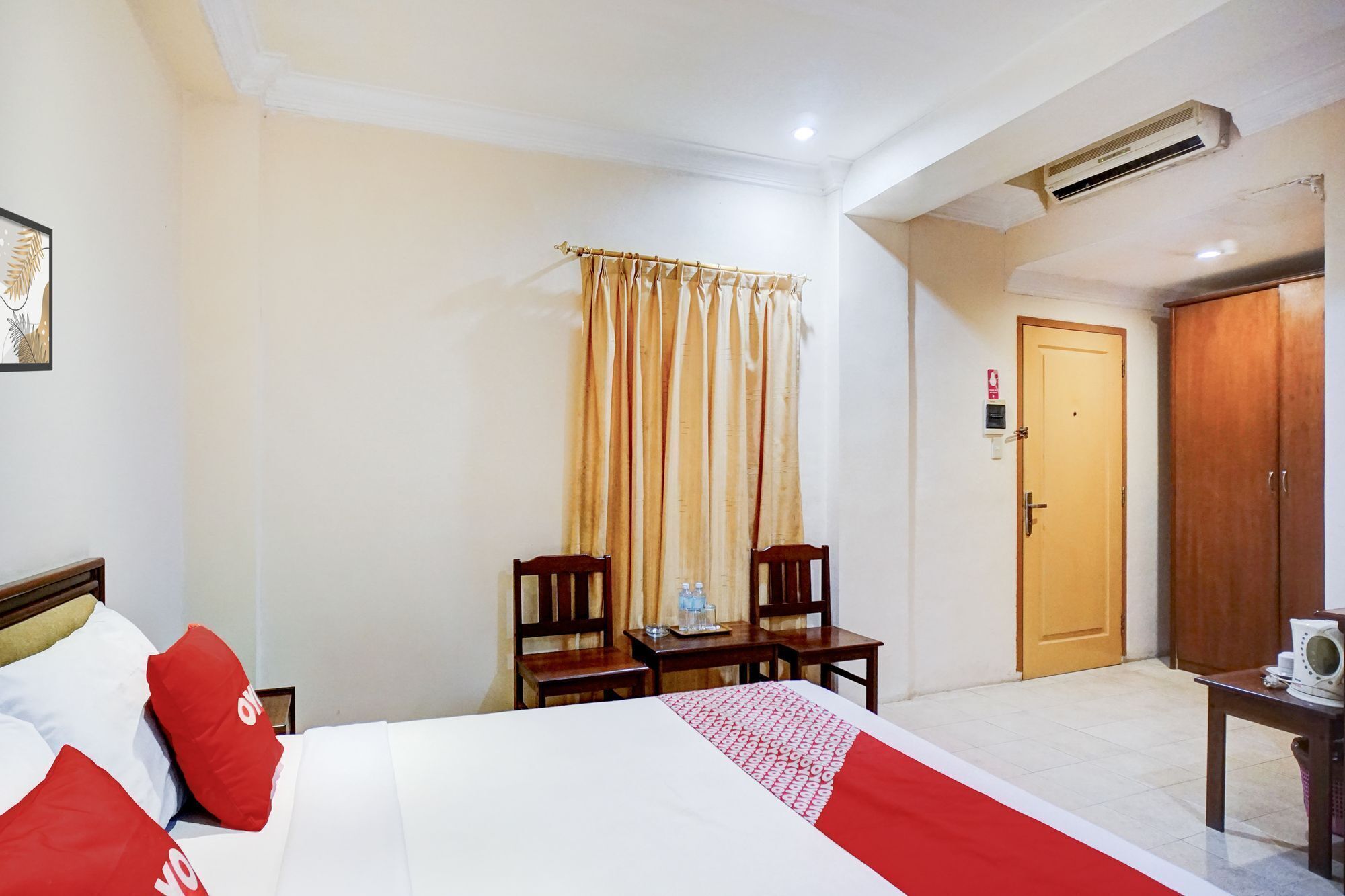 A hotel room with a bed, two chairs, a table, a door, and an air conditioning unit.