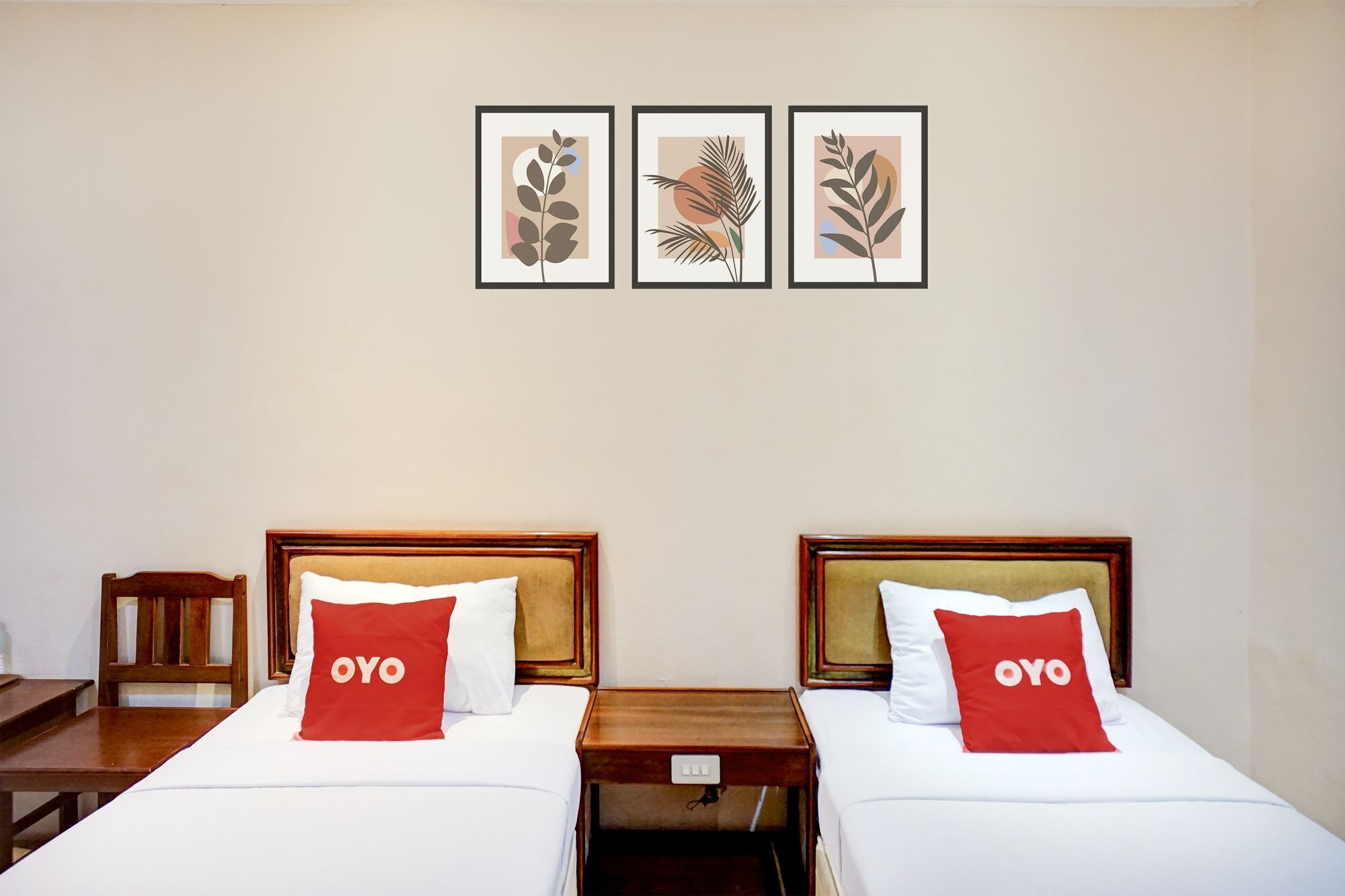 A hotel room with two beds, each with a red pillow labeled 'OYO,' a wooden nightstand, and a chair. There are three framed botanical prints on the wall.