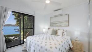 Un dormitorio con un ventanal grande, una cama con ropa de cama estampada, un cuadro sobre la cama y una mesita de noche pequeña con una lámpara.