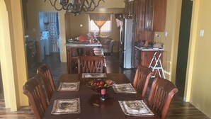 Un coin repas est aménagé autour d’une table en bois, dressée pour six personnes, et entouré de chaises dont les dossiers présentent des motifs complexes. Un lustre est suspendu au plafond, et la pièce est dotée d’un plancher en bois et de murs jaunes.