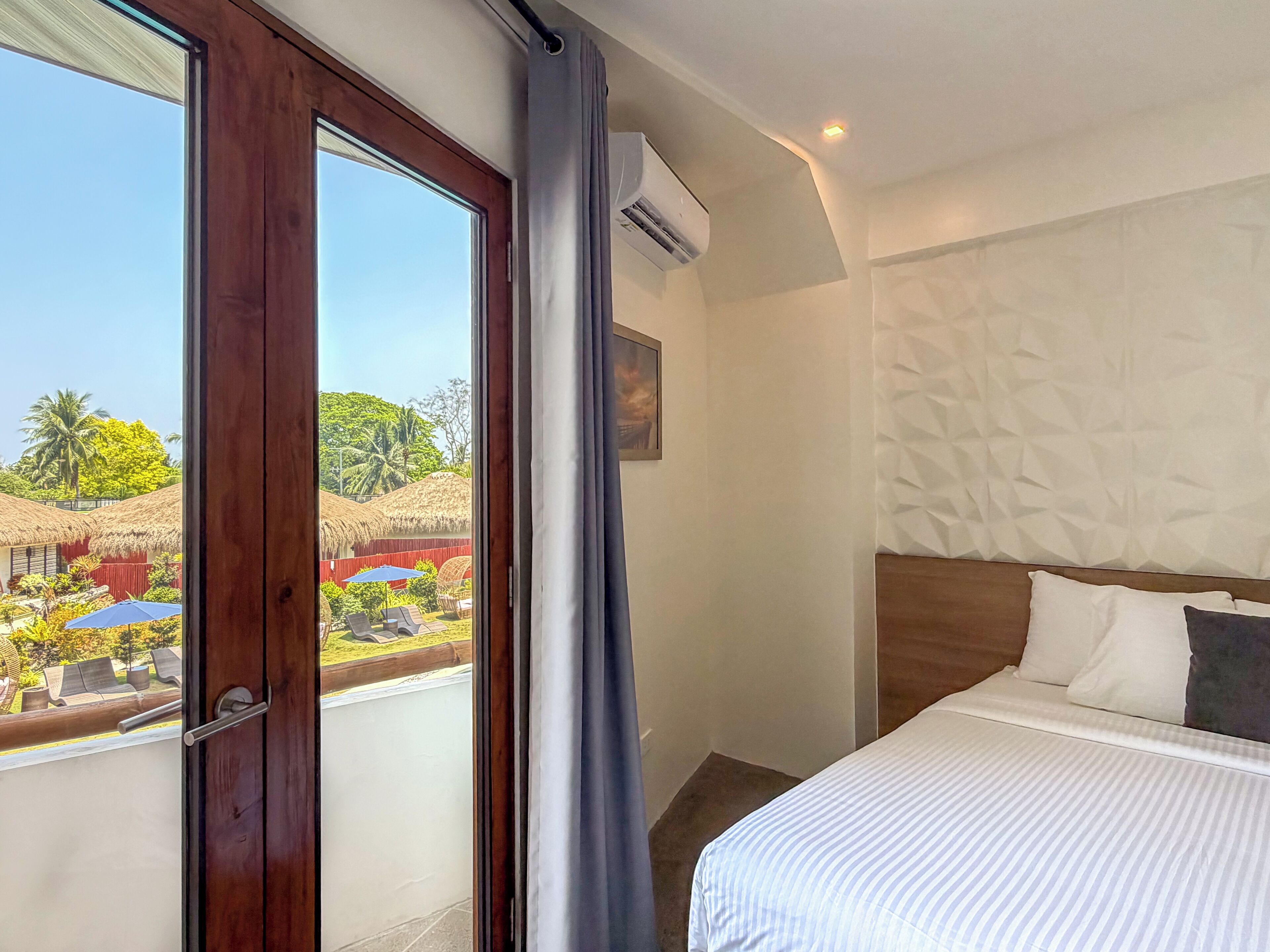A hotel room with a large window, a bed with white bedding, a wooden headboard, and an air conditioning unit.
