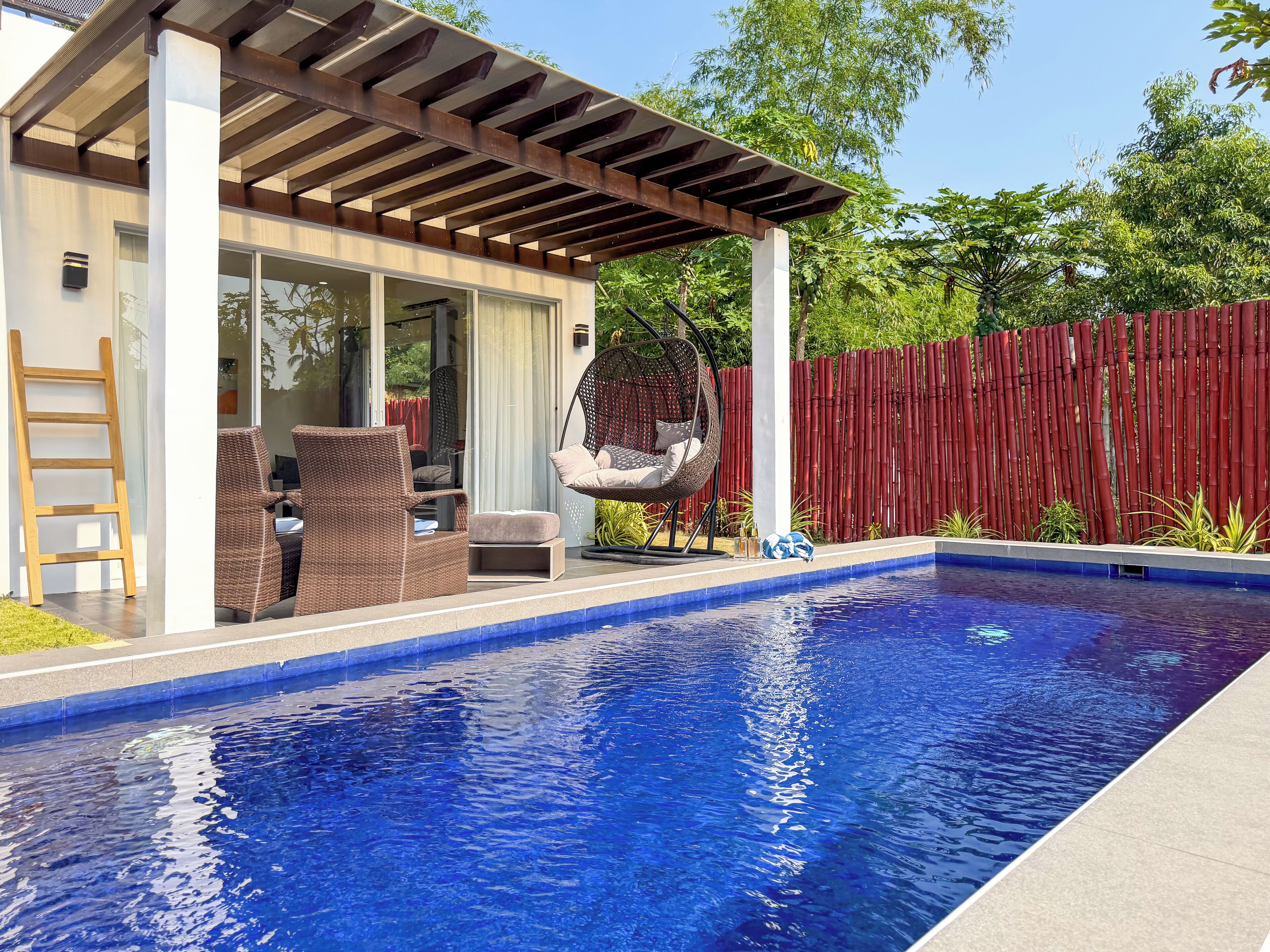 A swimming pool with a pergola, wicker furniture, and a hanging chair.